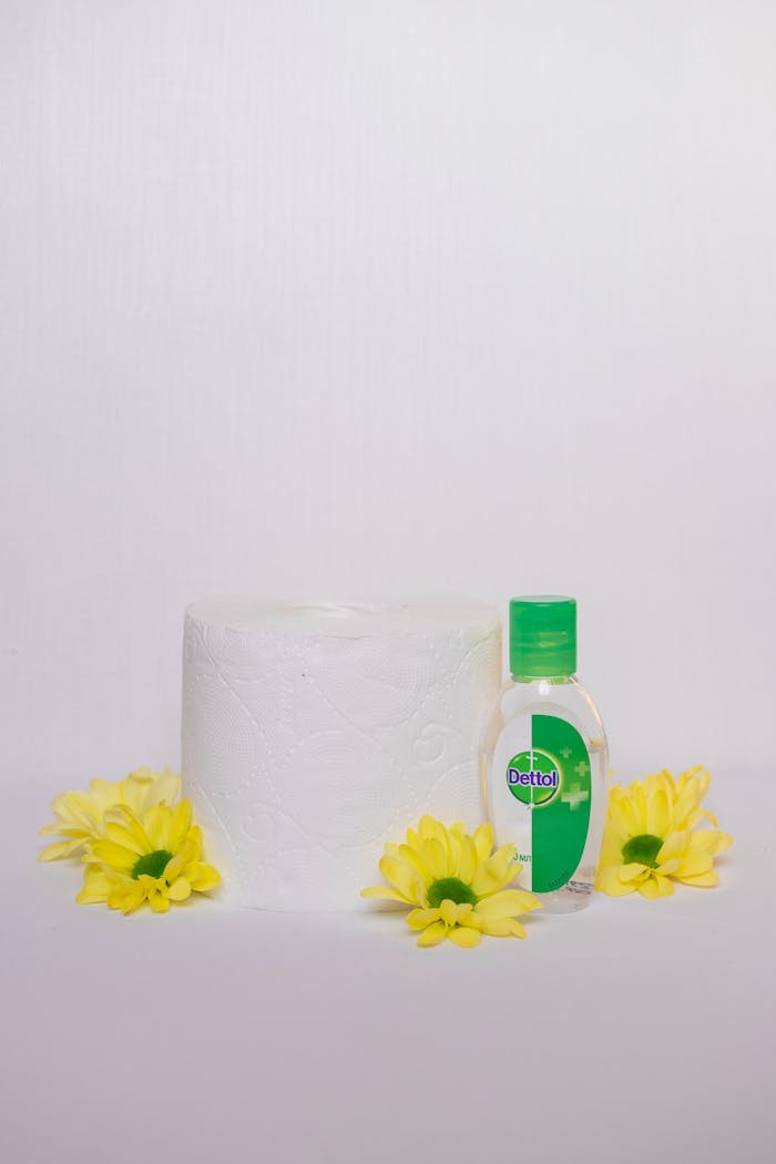 Tissue roll and hand sanitizer bottle surrounded by yellow flowers on a neutral background.